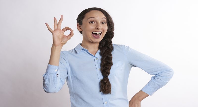 Motiv: Smilende ung kvinde i blå skjorte laver ok-tegn med fingrene