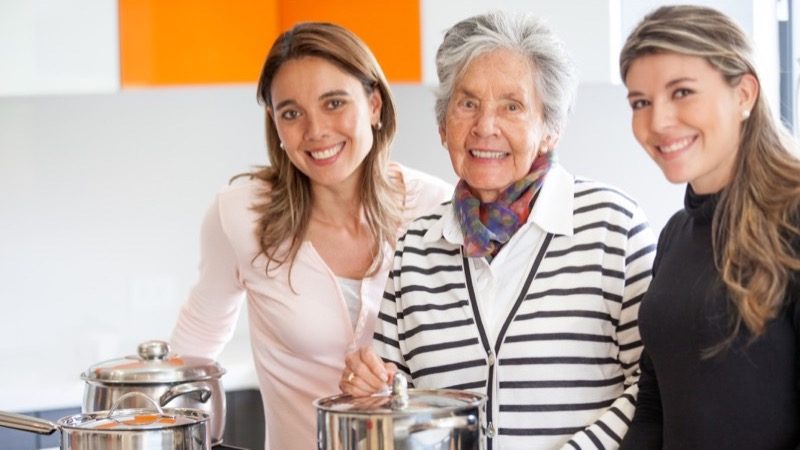 Motiv: Ældre kvinde i køkken flankeret af to yngre kvinder