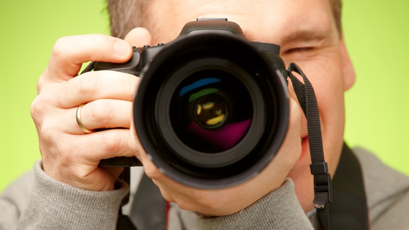 Nærbillede af en mand, der holder et kamera op foran ansigtet og tager et billede. Kameraets store objektiv fylder det meste af billedet, og man kan se refleksioner i linsen. Personen bærer en grå trøje og har en vielsesring på fingeren. Baggrunden er ensfarvet grøn og sløret.